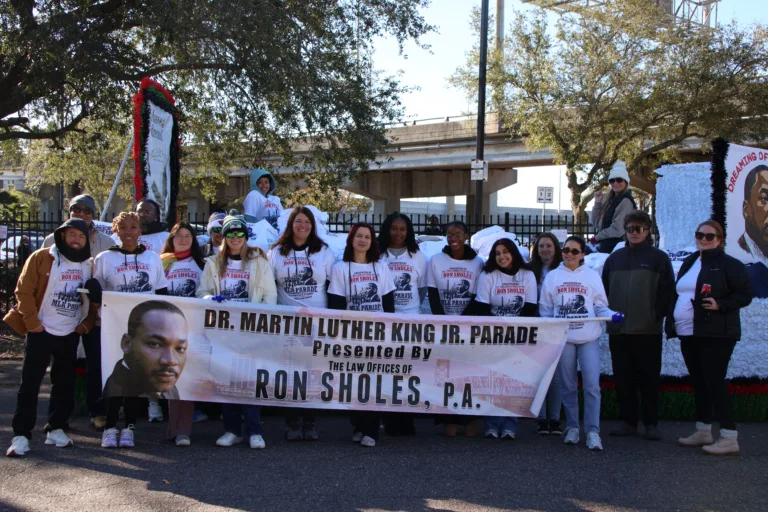 Volunteers and staff from the Law Offices of Ron Sholes, P.A. gather for a group photo at the Dr. Martin Luther King Jr. Parade. They are dressed in commemorative white shirts and holding a large event banner near a float decorated with Dr. King’s image.
