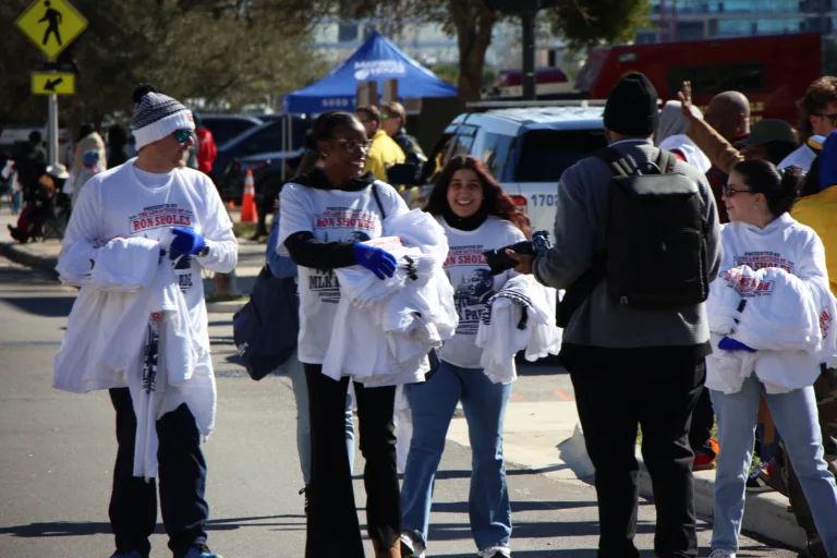 Smiling volunteers carrying stacks of white t-shirts to distribute during an MLK Jr. Day community event.