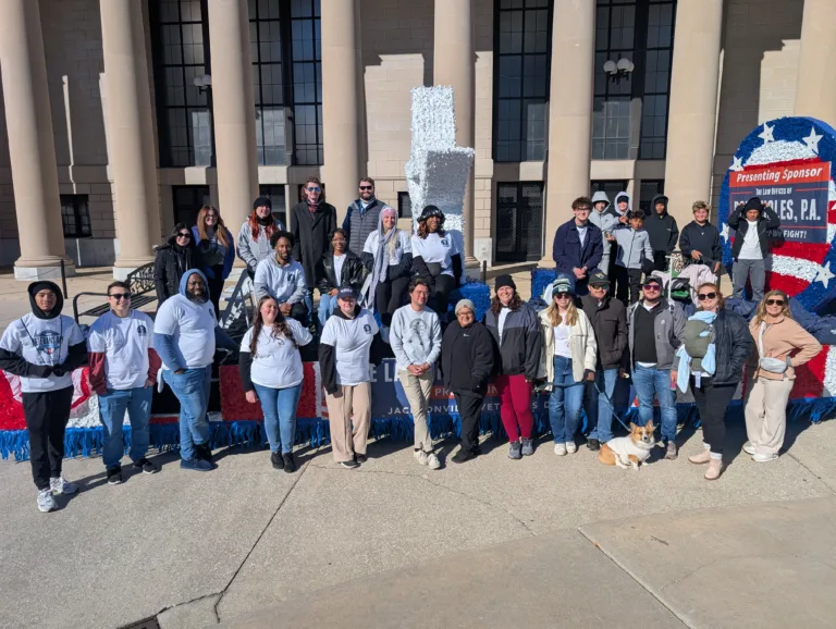 A large group of employees posing outdoors in front of a float decorated with red, white, and blue for Veterans Day. The group includes men and women of various ages, some wearing white shirts with the law firm’s logo. They are standing on a paved area in front of a building with tall columns. A sign on the float reads “Presenting Sponsor: The Law Offices of Ron Sholes, P.A
