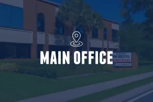 A wide-angle, low-light shot of a two-story brick professional building featuring large windows and palm trees. A white location pin icon and the text "MAIN OFFICE" are centered in the foreground. A sign for "The Law Offices of Ron Sholes, P.A." is visible on the lawn.