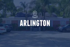 A daytime photo of a single-story brick building with white trim and a small portico entrance, flanked by palm trees. Several cars are parked in the lot in front. The image is overlaid with a location pin icon and the text "ARLINGTON."