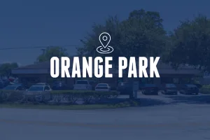 A horizontal view of a long, single-story brown building behind a manicured hedge and a parking lot filled with various cars. Large trees provide shade in the background. The image features a centered location pin icon and the text "ORANGE PARK."