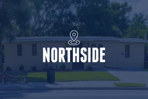 A front-facing shot of a tan, modern single-story building with narrow vertical windows and a flat roof. A green lawn and sidewalk are in the foreground. The image is overlaid with a location pin icon and the text "NORTHSIDE."