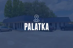 An angled shot of a light blue building with a dark blue metal mansard roof. The building is surrounded by a paved parking lot with several vehicles. A location pin icon and the text "PALATKA" are centered in the frame.