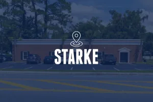A wide view of a red brick building with white decorative trim above the windows and doors. The building sits behind a parking lot with several trucks and SUVs. The image is overlaid with a location pin icon and the text "STARKE."
